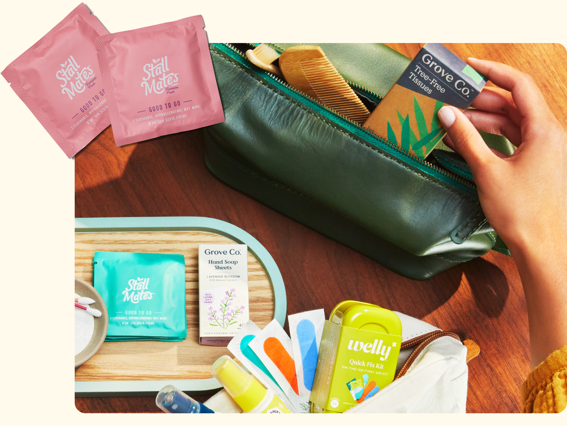 A hand reaches into a green bag containing toiletries, including Grove Co. soap. Nearby are Stall Mates wipes, nail files, a Welly first aid kit, a comb, and other personal care items on a tray and table.