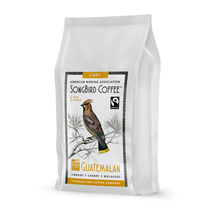 A white bag of Songbird Guatemalan Light Roast by Thanksgiving Coffee features a bird illustration and reads: American Birding Association, Light Roast, Guatemalan Bird Friendly Coffee, Vibrant, Cherry & Molasses. It includes the Fair Trade logo and is made in the Maya Highlands.