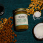 A jar of Big Spoon Roasters Toasted Coconut Almond Butter sits among toasted coconut flakes, a halved coconut, and sea salt on a dark green surface—a delicious, low-sugar nut butter.