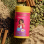 A jar of Diaspora Spice Co. Chai Spice is displayed amidst ground spices, whole cardamom pods, cinnamon sticks, and black peppercorns. The label features an illustration of a woman enjoying chai at a table with cookies, celebrating the single-origin ingredients that make this chai masala unique.