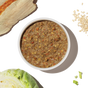 A bowl of The Honest Kitchen Dehydrated Whole Grain Turkey Dog Food sits on a white background, surrounded by raw chicken breast, oats, chopped cabbage, and green vegetables.