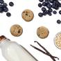 A pack of The Honest Kitchen Dog Treat Blueberry & Vanilla Goats Milk N Cookies is displayed with blueberry cookies, fresh blueberries, oats, and vanilla beans on a black background.