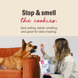 A woman sits on an orange couch with a large dog, holding The Honest Kitchen Dog Treat Peanut Butter & Honey Goats Milk N Cookies above its head. Text above reads, Stop & smell the cookies.