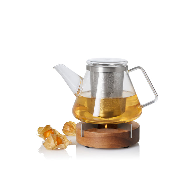 The AdHoc Borosilicate Glass Teapot with Stainless Steel Infuser and Candle Warmer sits on an acacia wood base filled with light amber tea, beside three physalis fruits, all set against a white background.