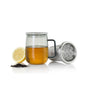 An AdHoc Borosilicate Glass Tea Mug with Stainless Steel Infuser sits on a white background, accompanied by a lemon half and loose large-leaf tea.