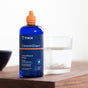 A blue bottle of Trace Minerals ConcenTrace stands on a wooden surface beside a glass of water, with a blurred bowl in the background.