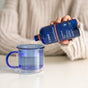 A person in a beige sweater pours ConcenTrace by Trace Minerals, a blue bottle of ionic trace minerals, into a clear mug of water on a white table.