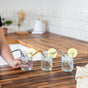 Three glasses of iced water with lemon slices feature EcoLunchBox's Turtleneck Stainless Steel Bendable Straws on a wooden countertop, with a hand holding the left glass. The background includes a tiled wall and kitchen items.