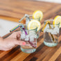 Three glasses of iced water with lemon slices and EcoLunchBox Turtleneck Stainless Steel Bendable Straws sit on a wooden surface. A hand holds one glass, featuring a rainbow-hued reusable straw from the set.
