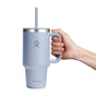 A hand holds a Hydro Flask Travel Tumbler, 32 oz, in powder blue with a leak-resistant lid and straw. The mug displays a logo of a stylized person with raised arms and has double-wall vacuum insulation to keep your drink at the ideal temperature against a plain white background.