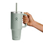 A hand holds a 40 oz green Hydro Flask Travel Tumbler with handle and straw, showcasing double-wall vacuum insulation, a stainless steel rim, and the Hydro Flask logo against a plain white backdrop.