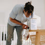 A person refills a reusable bottle with The Unscented Company Dish Soap Refill from a box labeled “Keeps your dishes and our waterways squeaky clean,” next to a wooden table and woven chair.