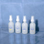 Four bottles of TUSHY Toilet Spray—Morning Zest, Afternoon Dip, Late Bloomer, and Evening Wood—sit on glossy blue tiles, powerful odor eliminators ready to banish bathroom smells with every spritz.