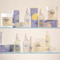 A display of TUSHY Toilet Spray in scents like Morning Zest, Evening Wood, Afternoon Dip, and Late Bloomer sits on two glass shelves with colorful boxes, designed to eliminate odors.