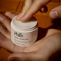 Someone uses their finger to scoop Phil's Tallow + Honey Balm from a white jar while holding it in their other hand. The grassfed tallow balm sits on a wooden surface.