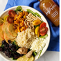 A bowl with rice, spinach, apples, cherry tomatoes, sweet potatoes, almonds, cranberries, cottage cheese, and grilled chicken sits beside a Kosterina Crushed Tangerine Vinegar bottle on a blue cloth.