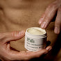 A person dips a finger into an open jar of Phil's Tattoo + Scar Recovery Balm, with a bare torso in the background, emphasizing tattoo aftercare and scar recovery.