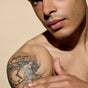 A man applies Phil's Tattoo + Scar Recovery Balm to his shoulder, which features a detailed clock tattoo and the words “ONLY TIME.” The background is soft beige.