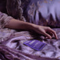 A hand rests on purple floral bedding beside a packet labeled Terra Herbal Nighttime Tea and a pack of Terra Intimate Toy Cleaning Wipes, individually wrapped in matching purple packaging, creating a calm and cozy vibe.
