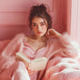A young woman with long brown hair relaxes on a plush pink sofa, reading about skincare while using Terra's Hydrating and Cleansing Micellar Makeup Remover Wipes for Face & Eyes—perfect for oily and acne-prone skin.
