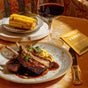A plate of lamb chops with sauce and pasta, served with red wine. In the background, grilled corn sits on a wooden board beside a yellow Terra Mess Free Multi-Purpose Wipes, Individually Wrapped packet.