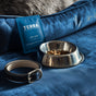 A metal pet food bowl with kibble, a black leather collar, and Terra Pet Grooming Wipes—fragrance free, gentle & hydrating, individually wrapped—sit on blue velvet near a gray pet bed, ideal for easy and natural pet care.