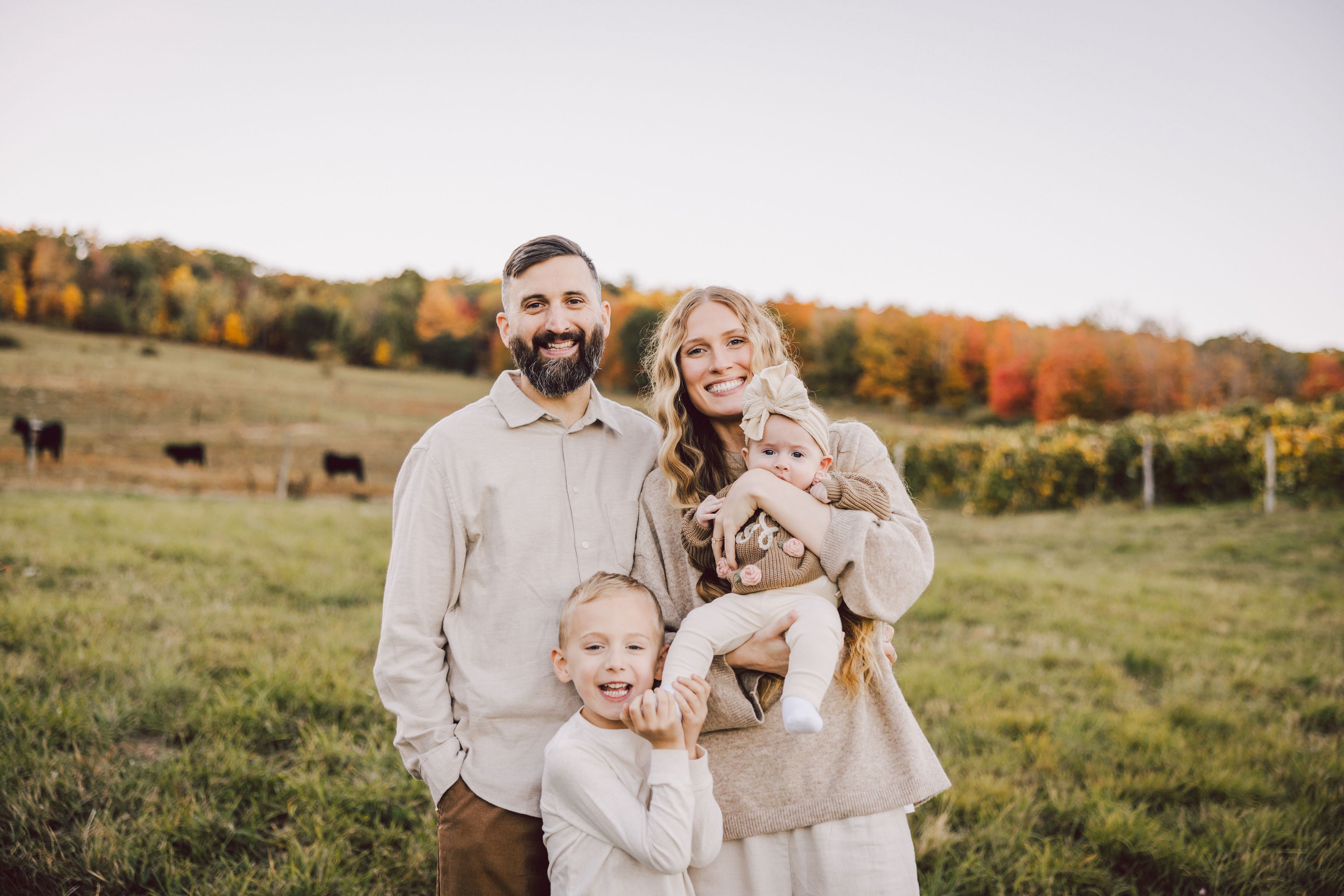 A smiling family of four stands in a grassy field with autumn trees in the background. The father has a beard, the mother holds a baby, and a young child stands in front, all dressed in light, neutral clothing.