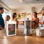 A moka pot, coffee mug, and a bag of Thanksgiving Coffee’s Holiday Blend sit on a kitchen counter as four people chat and smile in a warmly lit, festive kitchen.