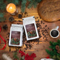 A cozy holiday scene with two bags of Thanksgiving Coffee’s Holiday Blend, a cup of black coffee, bread, a lit candle, pinecones, red berries, and evergreen branches arranged on a wooden table.