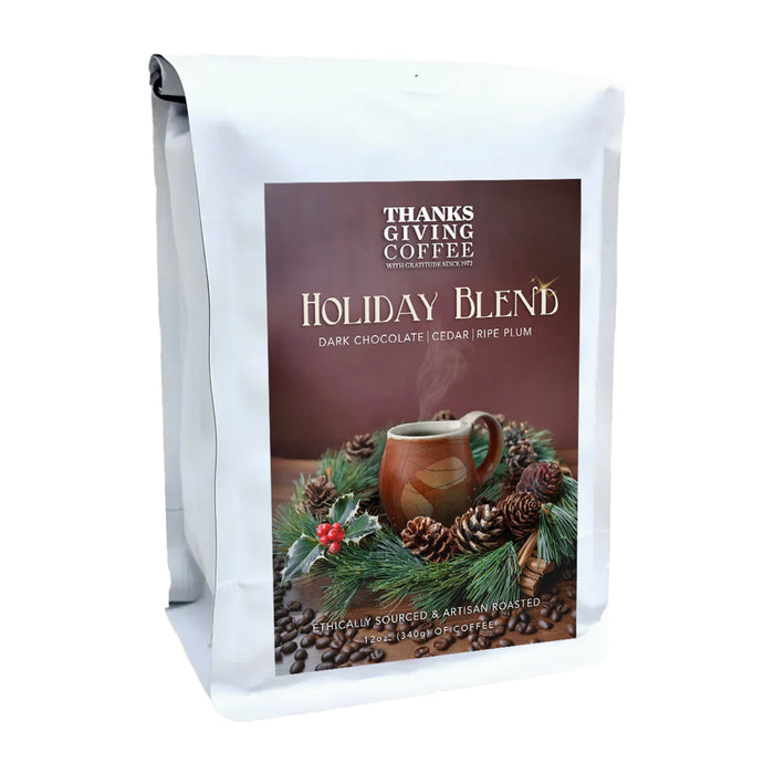 Thanksgiving Coffee’s Holiday Blend comes in a white bag with a festive mug, pine branches, pinecones, and coffee beans. The label notes rich flavors of dark chocolate, cedar, and ripe plum.