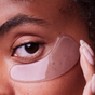A close-up shows a person applying The Good Patch De-Puff Hydrogel Undereye Patch, gently touching it to highlight a soothing skincare routine for under-eye care.