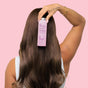 A person with long, smooth brown hair holds The Powder Shampoo’s Nourishing Repair Conditioner pink spray bottle against their head, showcasing tattoos on a raised arm against a light pink background, caring for damaged hair.