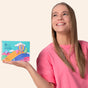 A smiling woman in a pink shirt holds a colorful Soar to New Heights box from The Powder Shampoo, featuring Cappadocia hot air balloons, a rainbow, and clouds. She looks up and to the side.
