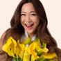 A smiling woman with long brown hair holds a bouquet of yellow calla lilies and a yellow bottle of The Powder Shampoo Invigorating Powder Shampoo for Thinning & Ageing Hair against a light background.