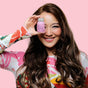 A smiling woman with long, wavy brown hair restored from damage holds a small pink bottle of The Powder Shampoo’s Nourishing Repair Conditioner to one eye. She wears a colorful long-sleeve top against a plain pink background.