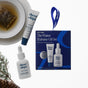 The Winter Hydration Gift Set containing a lip treatment tube and a dropper bottle of serum, with a loose lip treatment, a dropper bottle, a tea bag in a saucer, and a pine sprig nearby.