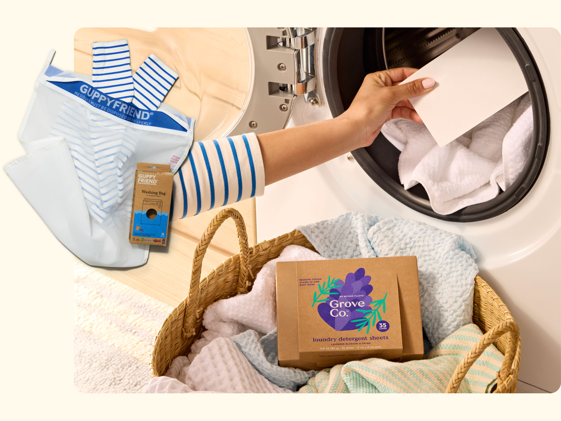A hand places a sheet into a front-load washing machine filled with laundry. Inset shows a Guppyfriend washing bag. A wicker basket with towels and a box of Grove Co. laundry detergent sheets is nearby.