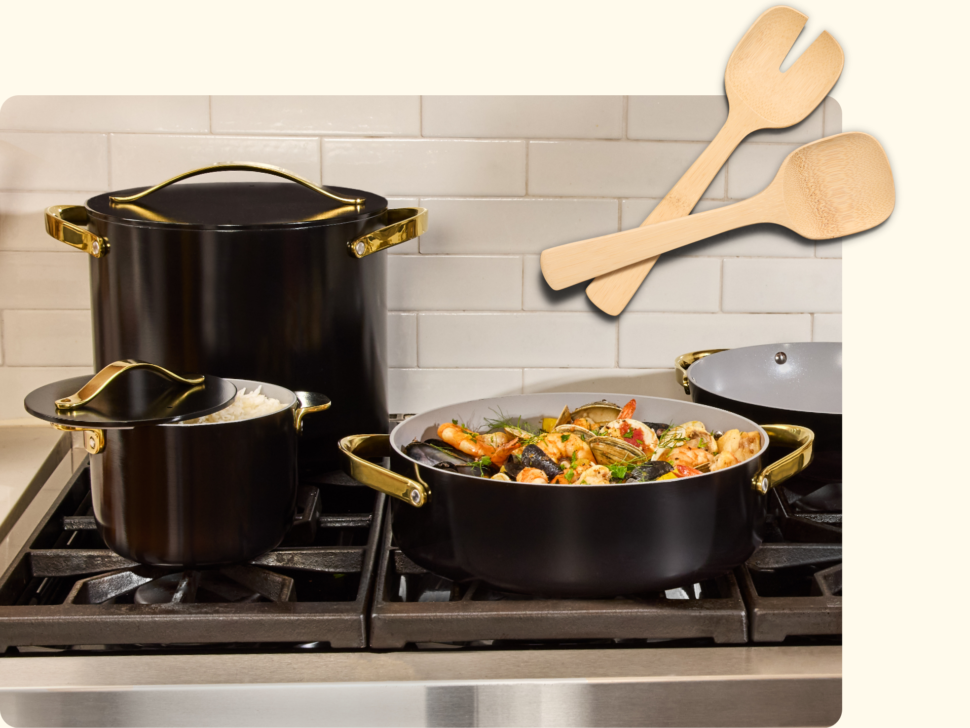 Three black pots with gold handles on a stovetop, one filled with cooked rice, another with seafood and vegetables. Two wooden utensils are placed beside the pots. White subway tile backsplash.