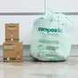A large green Compostic 13 Gallon Home Compostable Trash Bag sits on a concrete floor beside two boxes labeled Compostic 100% compostable kitchen bags, with white cabinets visible in the background.