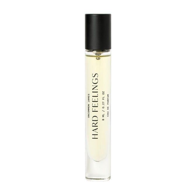 A clear glass 8 mL vial with a black cap holds pale yellow Hard Feelings Eau de Parfum by Uncommon Beauty. The bold label hints at an alluring sandalwood aroma and notes Eau de Parfum along with 0.27 fl oz in smaller text.