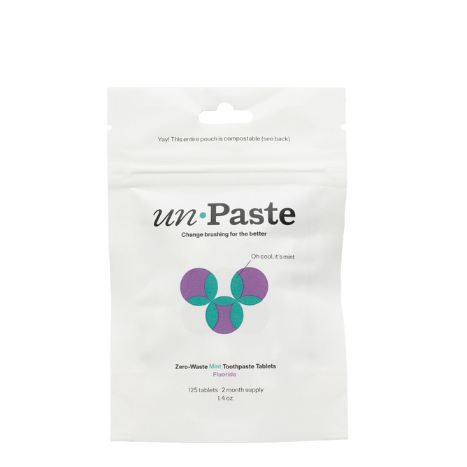 A white pouch labeled Unpaste Fluoride Mint Toothpaste Tablet contains natural, zero-waste toothpaste tablets. The plastic-free, compostable packaging features a green and purple circular design near the center.