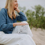 A smiling woman in a blue shirt and white pants sits outdoors on sand, holding a box of The Unscented Company Dishwasher Tabs. Green plants are in the background.