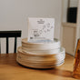 A box of The Unscented Company Dishwasher Tabs is placed on a stack of white plates on a wooden table, with a chair and other kitchen items in the background.