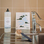 A bottle of The Unscented Company Dish Soap, a dish brush, and a scrubber sit on a kitchen counter beside a modern faucet and a decorative card with mountain and river art, all set against a tiled wall.