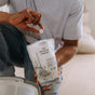 A person holds a Floor Cleaning Tab from The Unscented Company above an open bag with mop graphics. The eco-friendly tabs are ready to deep clean floors, with a bucket seen in the foreground.