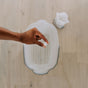 A hand holds a Floor Cleaning Tab by The Unscented Company above an empty white basin on a light wood floor, with a rolled-up white cloth in the background.