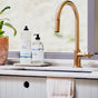A modern kitchen sink with a gold faucet, a potted plant, and glass bottles of The Unscented Company Hand Soap and dish soap on the counter by the window. A towel hangs over the white sink front.