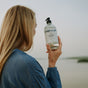 A person with long blonde hair in a blue shirt holds up a clear pump bottle of The Unscented Company Hand Soap while standing outdoors by a body of water.