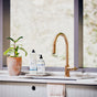 A modern kitchen features a white sink with a towel draped over the edge, a gold faucet, a potted plant in a terracotta pot, and The Unscented Company Hand Soap in glass dispensers on the countertop before large windows.