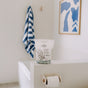A bathroom with a white bathtub, a striped blue and white towel, abstract blue and white artwork, toilet paper on a holder, and The Unscented Company’s Toilet Bowl Cleaning Tabs on the tub’s edge for sustainable hygiene.